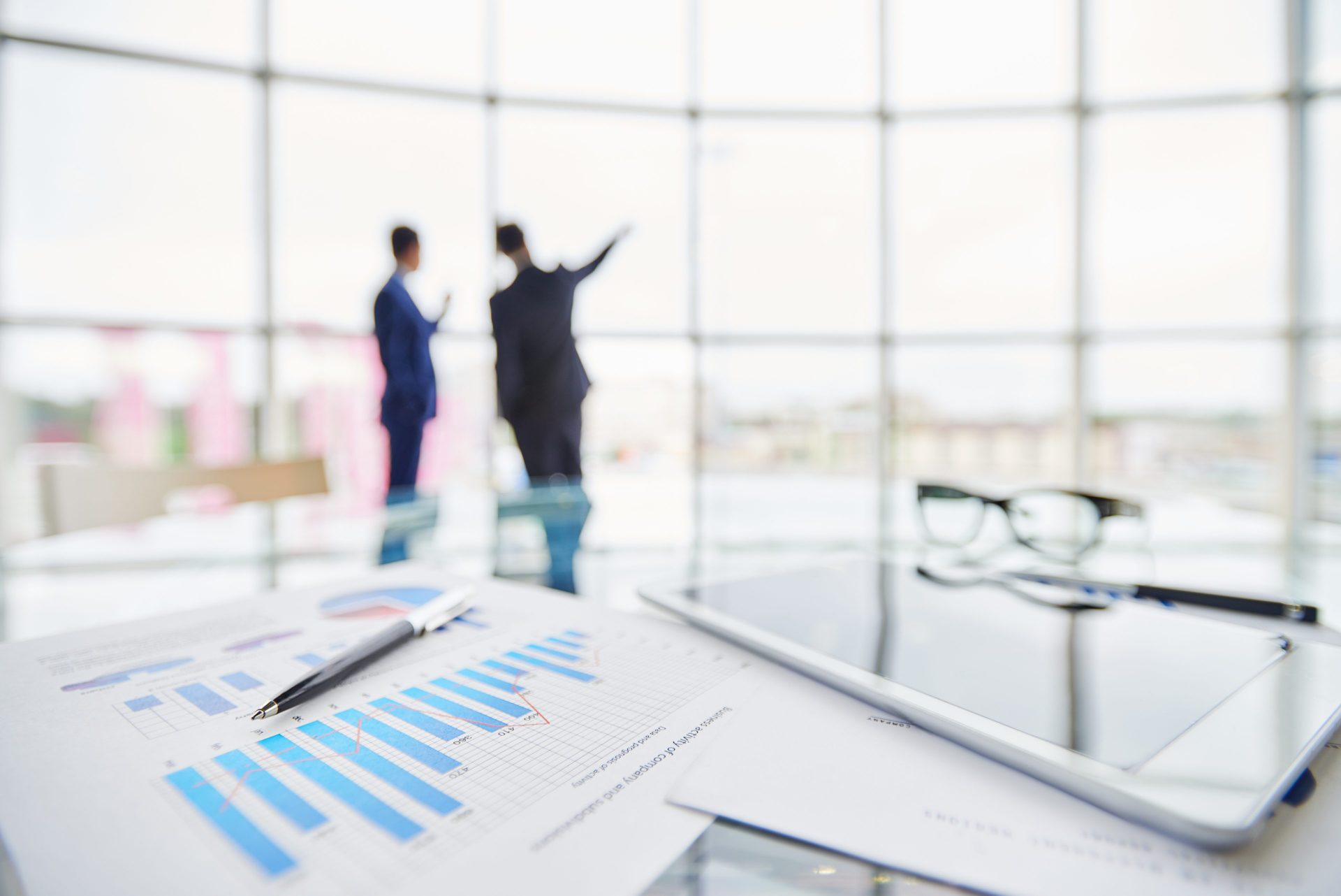 In focus are bar and line charts with touchpads and pens, out of focus are men speaking and pointing out the window.