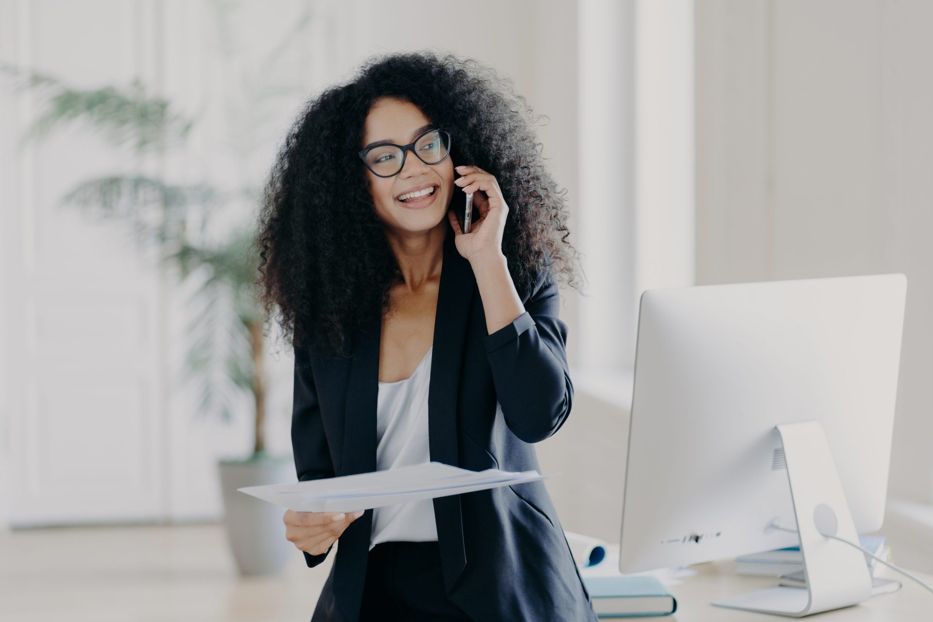 Women on her cell phone in an office next to her computer. Talking on her phone while smiling